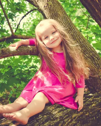 A blond girl, age 5, wearing a pink dress, happily sits on a large branch in a tree.