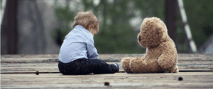 A toddler sits outside on cement, chatting with his favorite friend. A Teddy bear sits close by, listening.