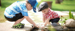 Two boys wearing ball caps lean over a small pond playing in the water.