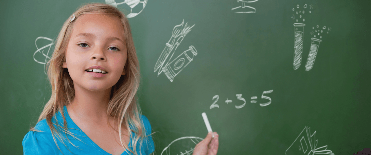 A young girl faces forward while holding chalk in her hands as she stands in front of a green chalkboard with various images and math problems written on it.