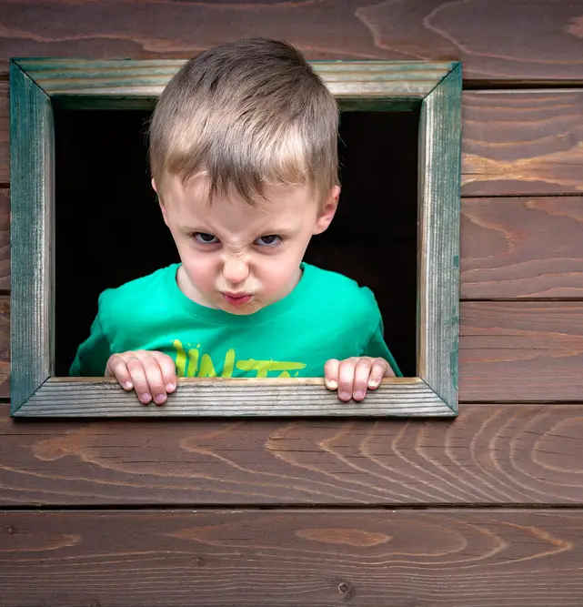 A little boy of four years sticks his head out of a playhouse window with an annoyed expression on his face.