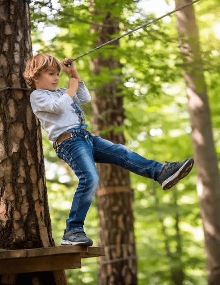 A young boy stands on a platform in a forested area, holding onto a rope and taking a step off the platform.