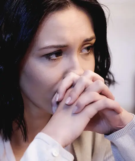 A woman with shoulder length brown hair sits with her hands to her mouth, deep in thought.