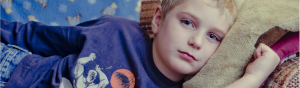 A boy of about 9 years old wearing a blue shirt lays on a couch staring at the camera expressionless.